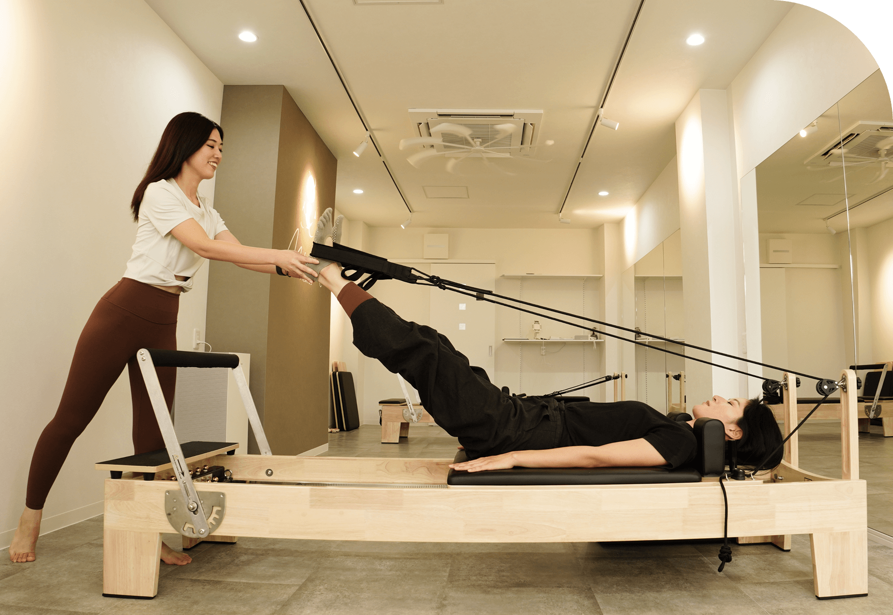 仰向けでピラティスマシンを使い、脚のトレーニングを行う女性とサポートするトレーナー
